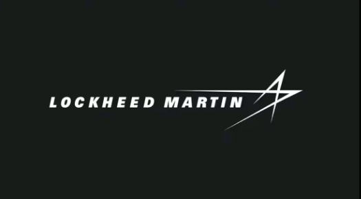 The image displays the logo of Lockheed Martin, a renowned aerospace and defence company. The logo features the company's name, "LOCKHEED MARTIN," in bold, white capital letters set against a black background. To the right of the text, there is a distinctive stylized emblem resembling a star or an arrowhead, also in white. The overall design conveys a sense of precision, innovation, and strength, which are qualities often associated with the company's work in the aerospace and defence industries.